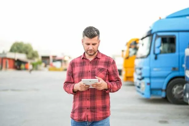 A fleet owner is in the lot of his business with trucks parked behind him as he reviews the analytics from his fuel card from the dashboard on his tablet.