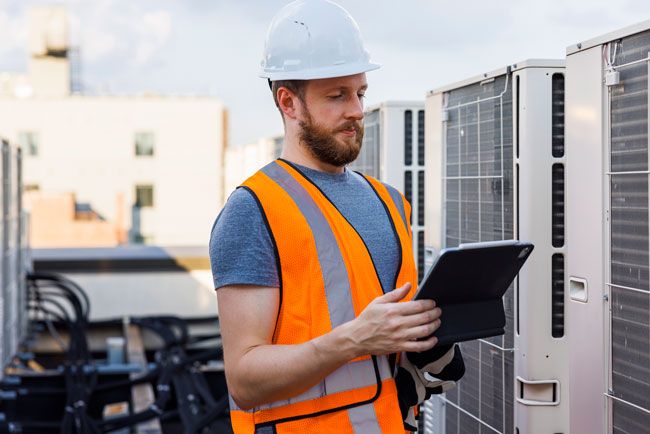 An HVAC contractor at a worksite wears a hard hat and reviews spending on a tablet.
